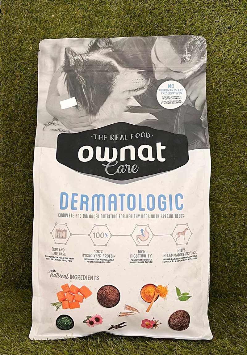 Bolsa de comida para perros Ownat Dermatologic sobre césped, envase blanco con texto negro y azul e iconos de ingredientes naranjas.