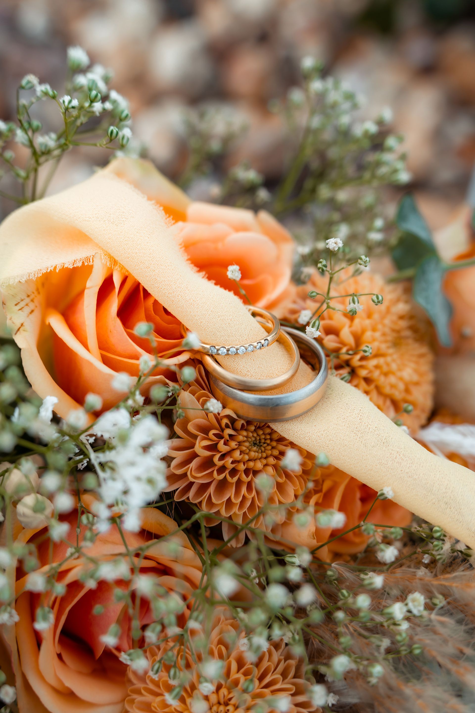 Eheringe liegen auf einem pfirsichfarbenen Band zwischen orangefarbenen Rosen und Schleierkrautblüten.