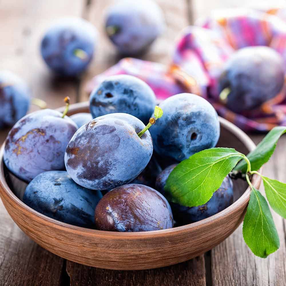 Eine Schale mit Pflaumen und grünen Blättern auf einem Holztisch.