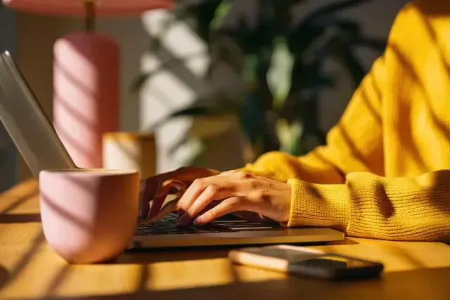Una persona con un suéter amarillo teclea en una computadora portátil junto a una