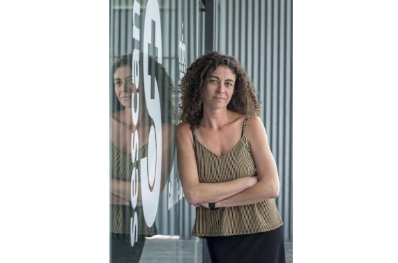 Una persona con cabello rizado, vestida con una blusa marrón estampada y una falda negra, permanece de pie con los 