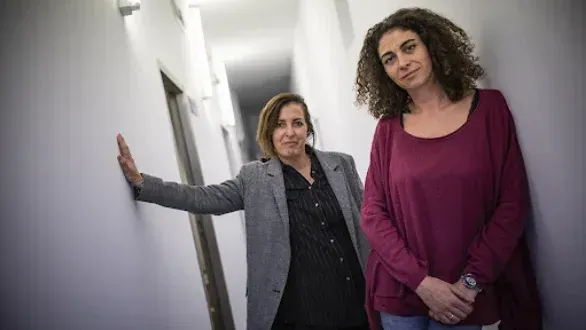 Dos personas se encuentran de pie en un pasillo estrecho y blanco; una está 