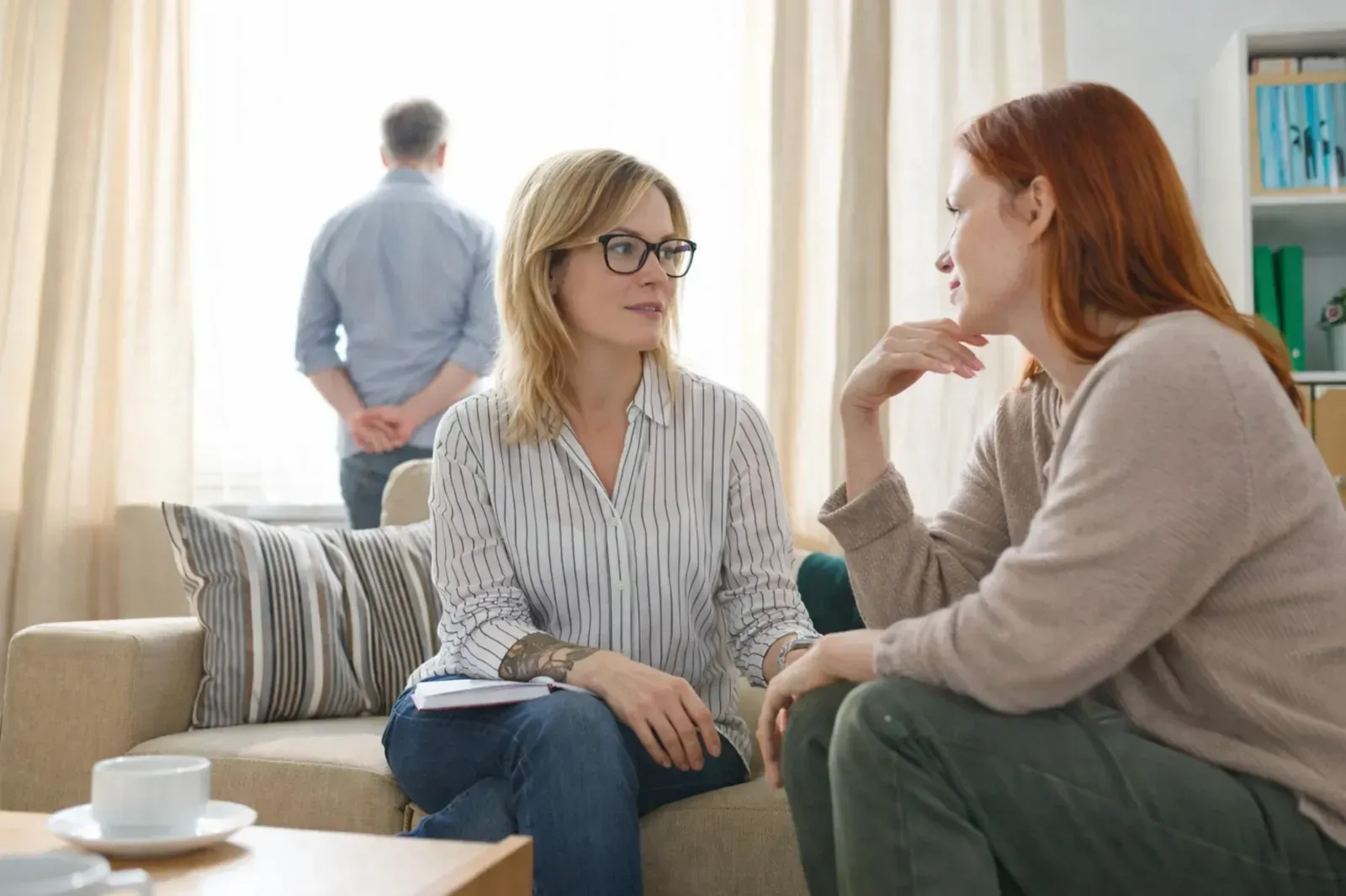 Dos personas conversan sentadas en un sofá, mientras una tercera persona