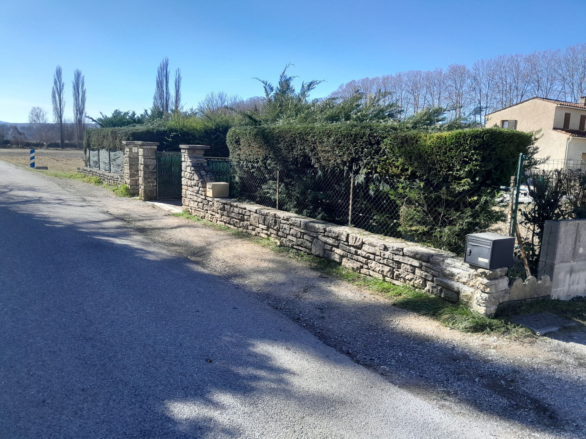 Clôture en pierre et haie le long d'un chemin de gravier devant une maison par une journée ensoleillée.