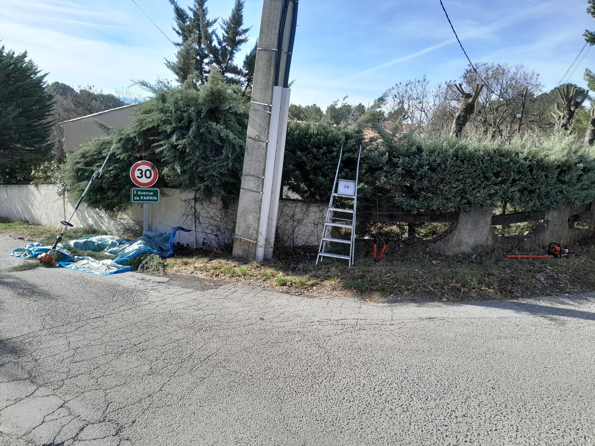 Échelle contre une haie taillée à côté d'un poteau électrique, bord de route avec des débris et un panneau de 30 km/h.