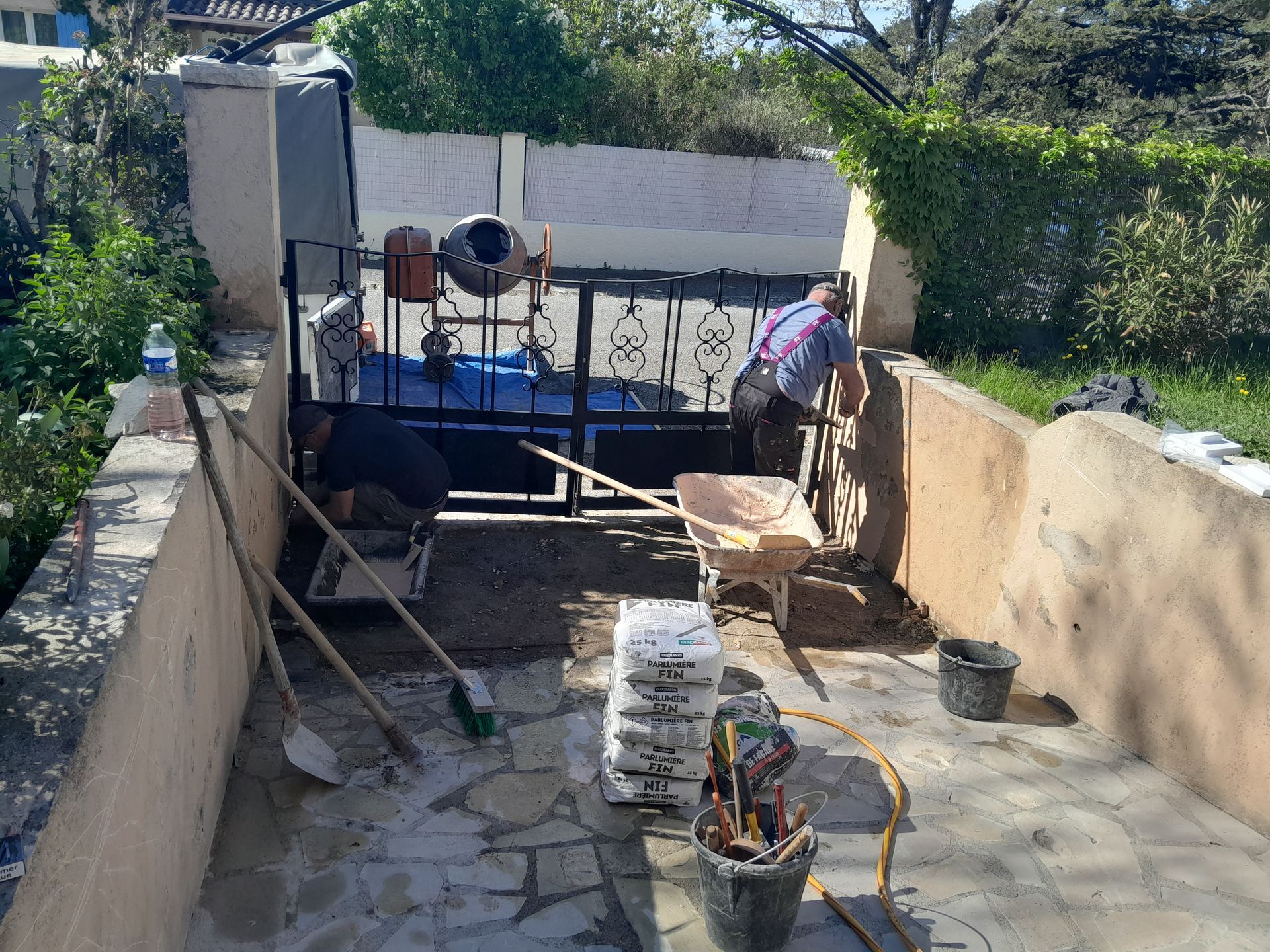 Des ouvriers travaillent dans un espace extérieur clos. Ils sont équipés d'outils, d'une bétonnière et d'une brouette.