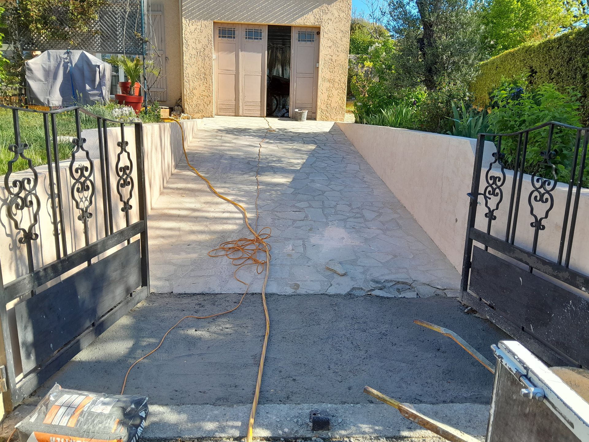 Allée ouverte en béton menant au garage, flanquée de murs et d'un portail en fer forgé. Matériaux de construction présents.