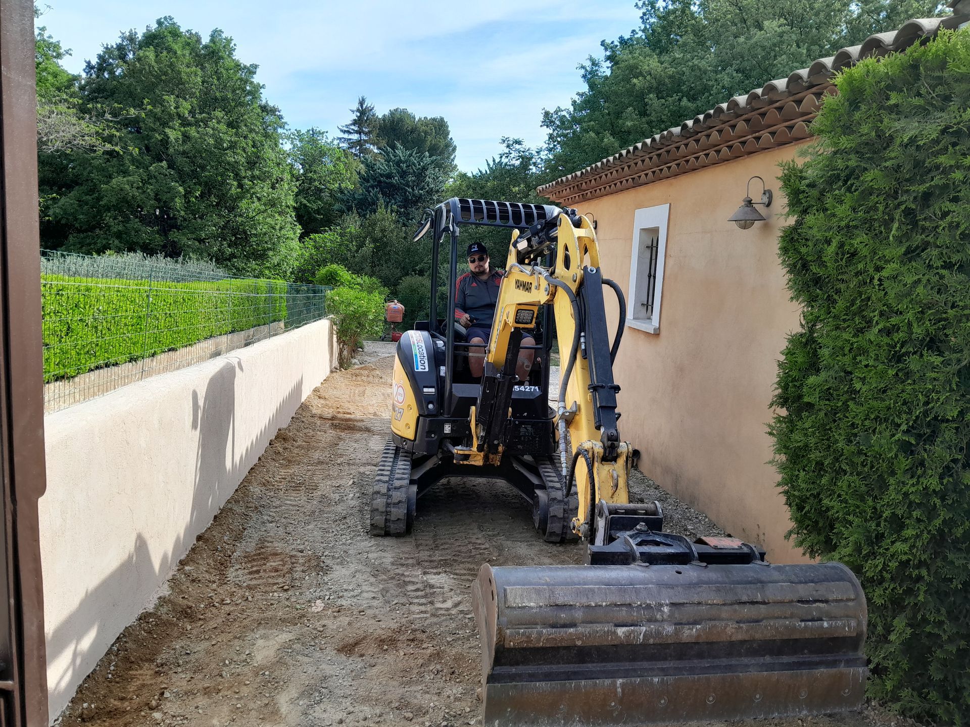 Un homme conduit une excavatrice jaune dans un chemin étroit à côté d'un bâtiment beige et d'une haie verte.
