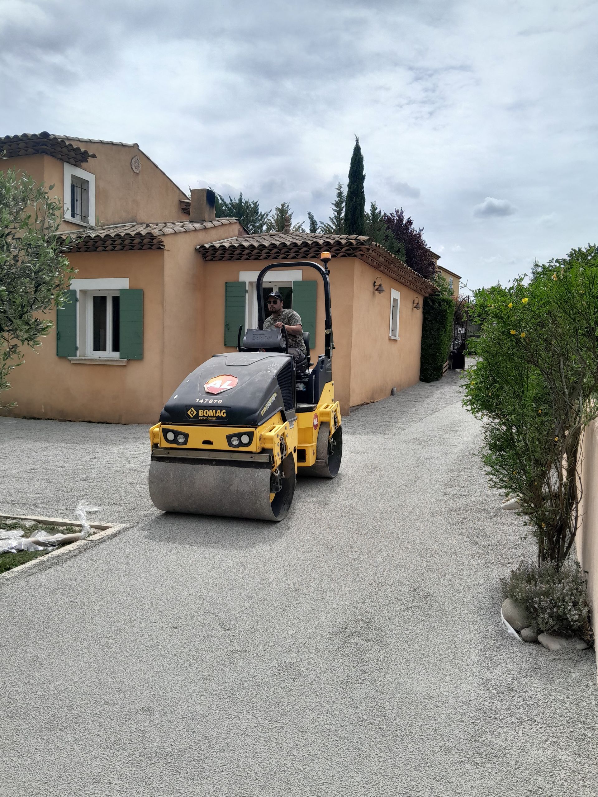 Un ouvrier du bâtiment utilise un rouleau compresseur jaune pour compacter une allée de gravier près d'une maison marron clair.