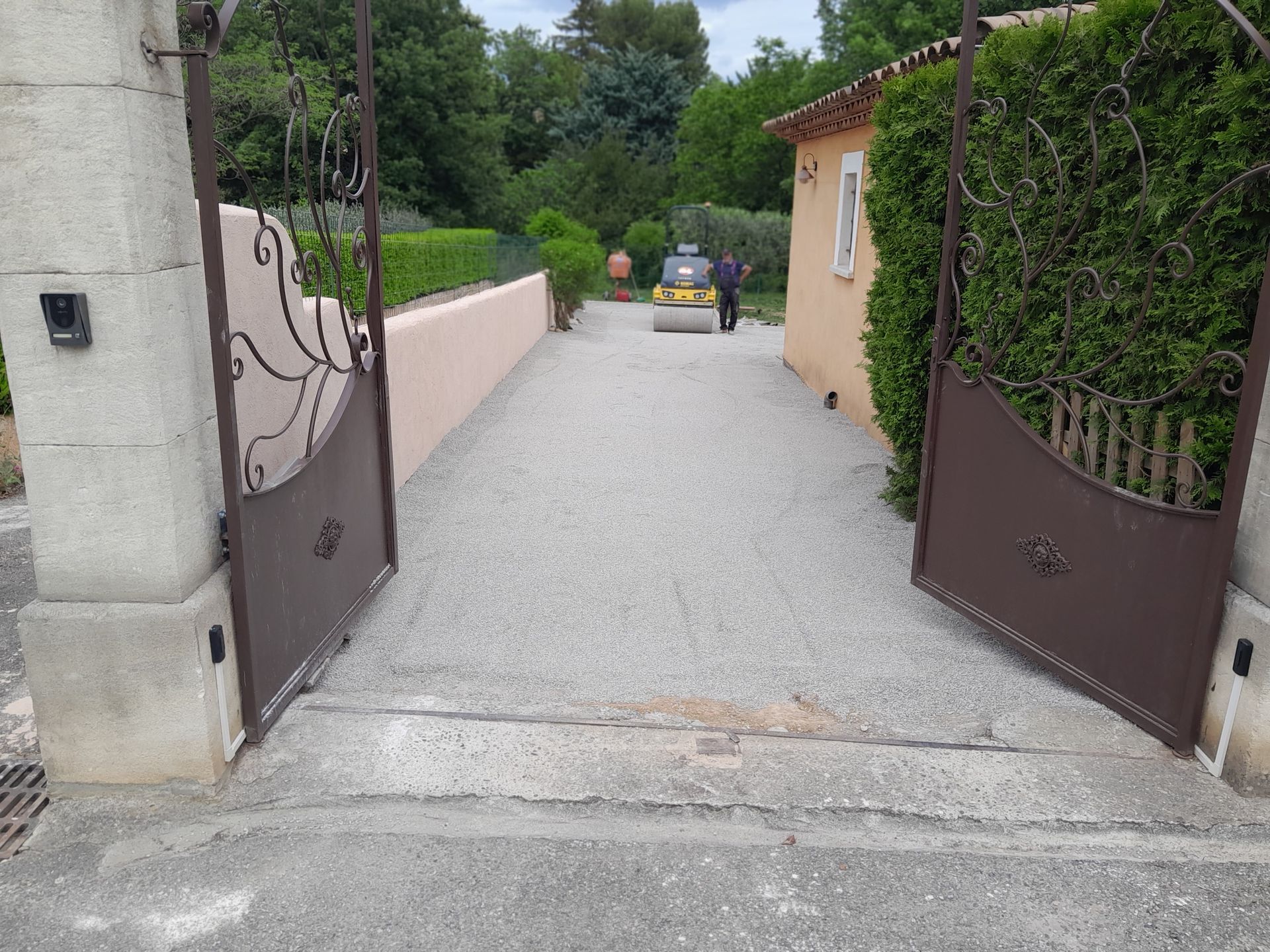 Allée de gravier nivelée par une machine, entre un portail en pierre et une maison. Arbres en arrière-plan.