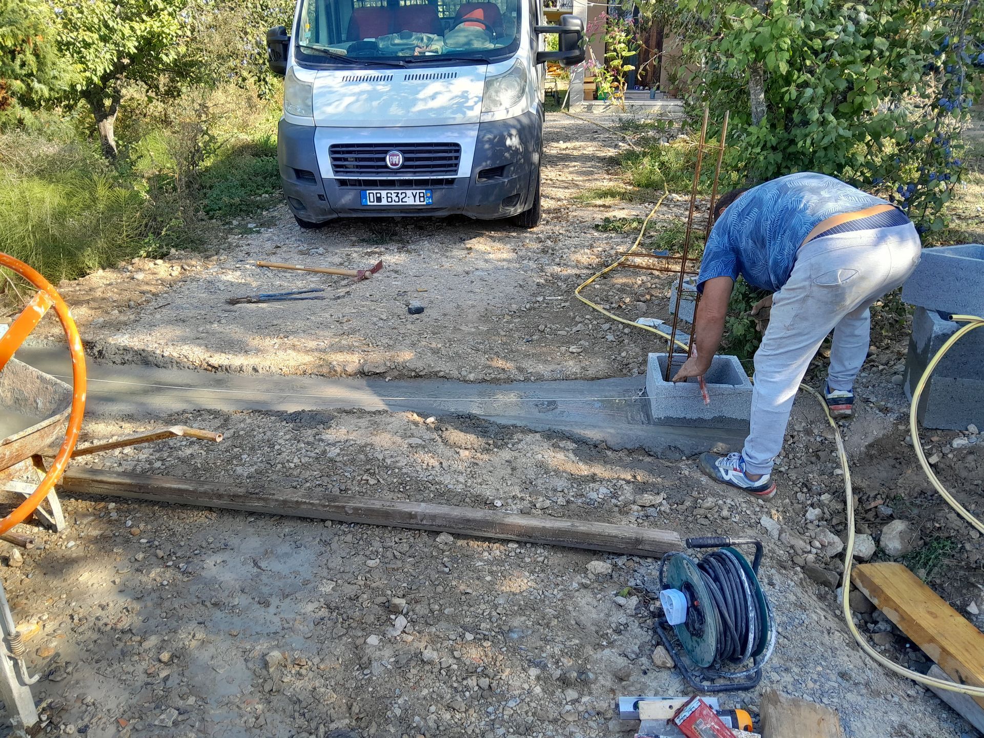 Un homme pose des parpaings sur du ciment frais près d'une camionnette. Scène de construction en extérieur avec des outils.