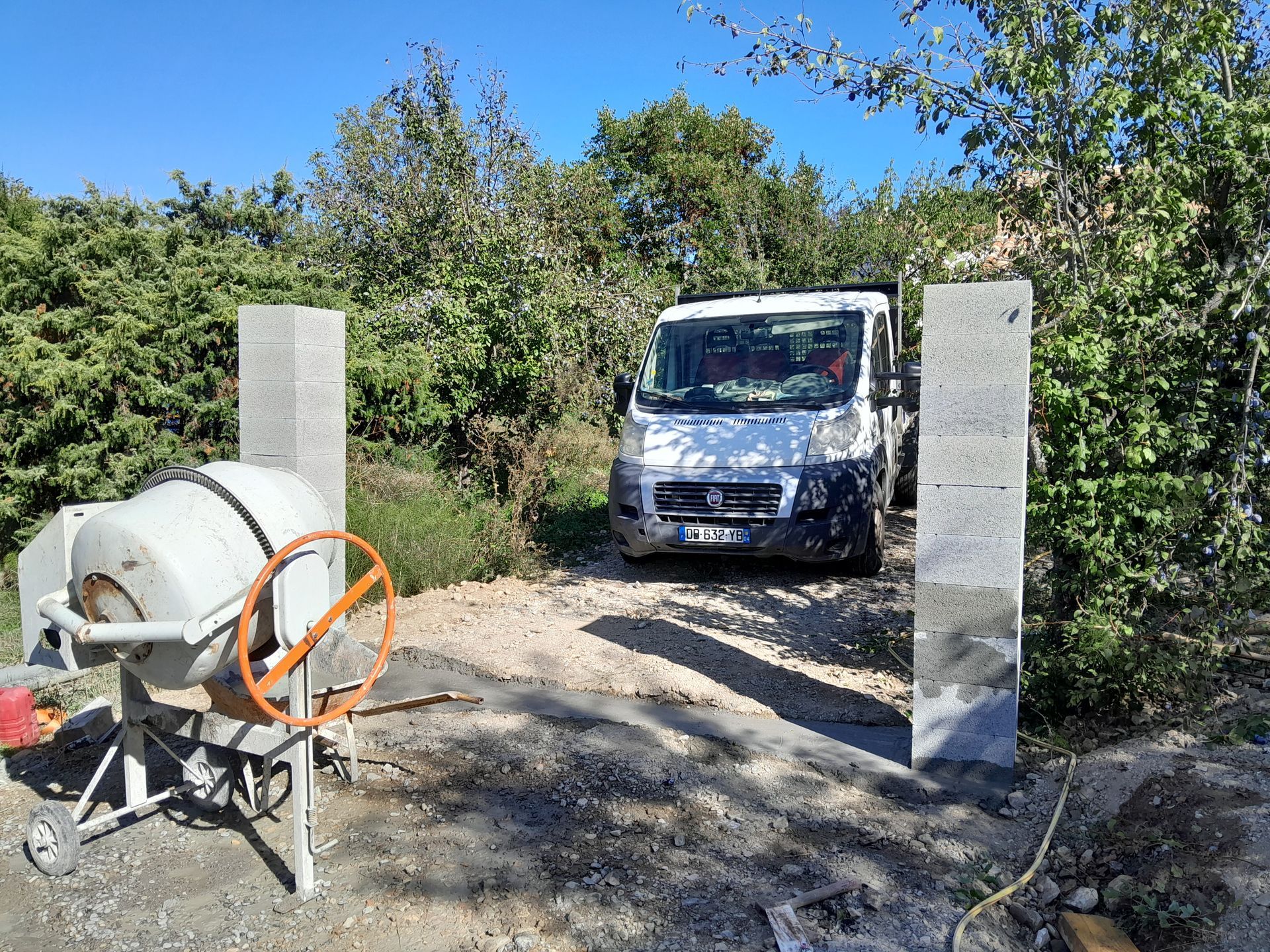 Chantier : une bétonnière à côté de colonnes de pierre, une camionnette blanche entre elles, des arbres en arrière-plan.