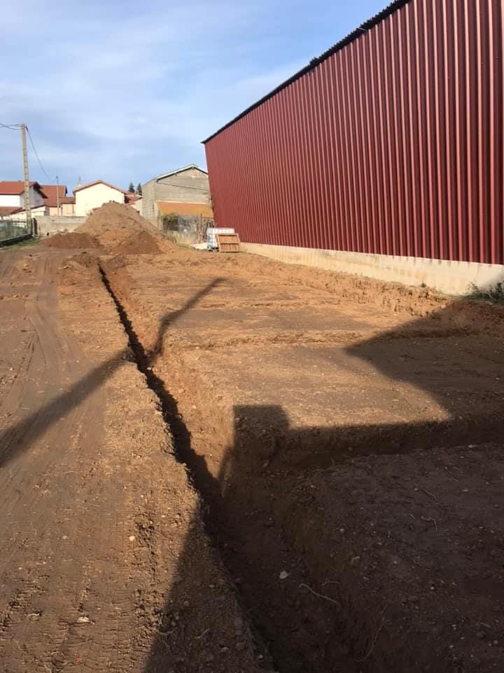 Une tranchée creusée dans la terre, le long d'un bâtiment métallique rouge, sous un ciel bleu.