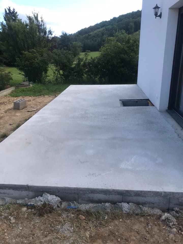 Terrasse en béton jouxtant une maison blanche, avec une trappe d'évacuation. Une colline verdoyante se dessine à l'arrière-plan.