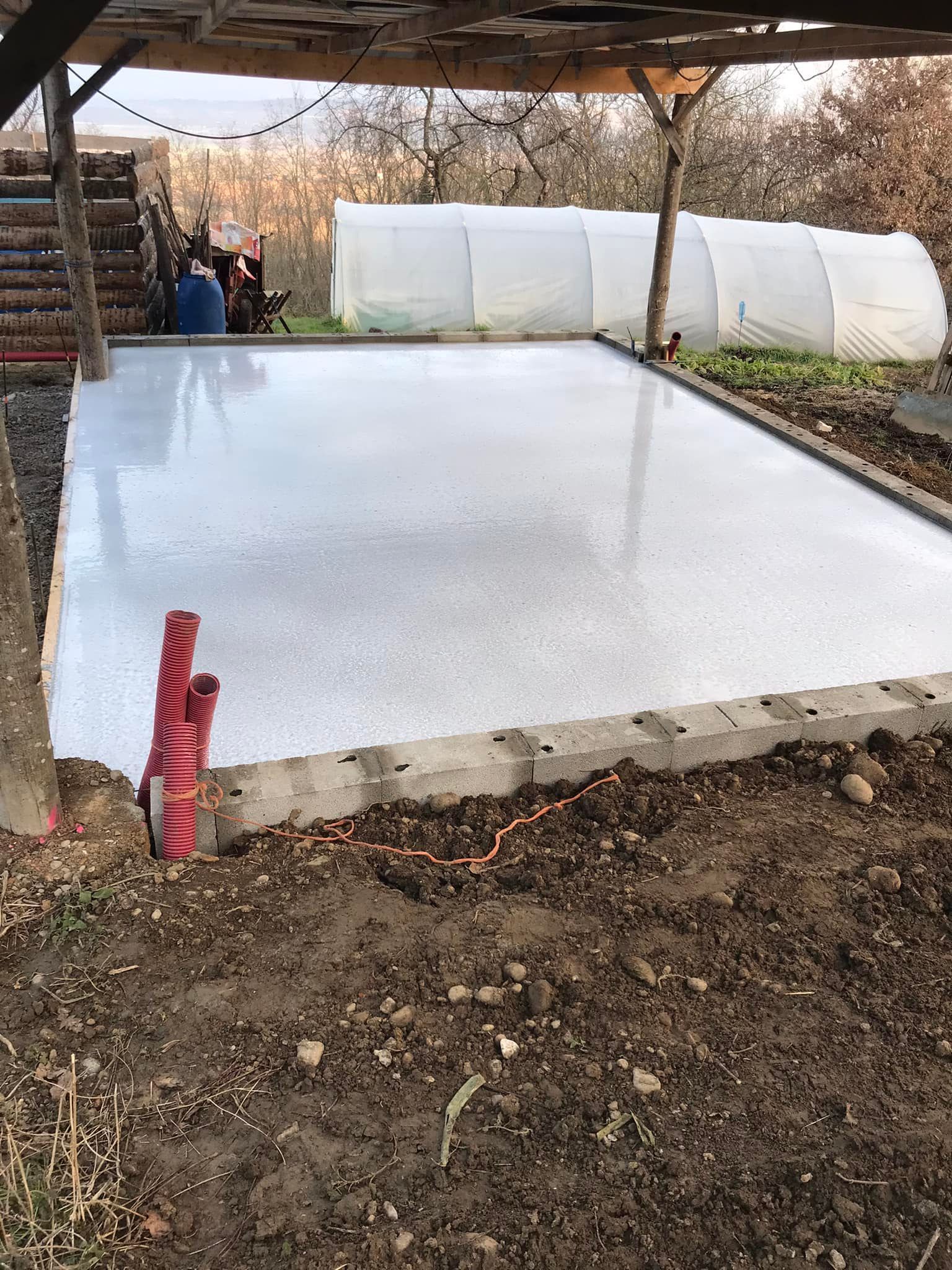 Terrasse en béton fraîchement coulé, entourée de terre, près d'une serre et d'une structure en bois.