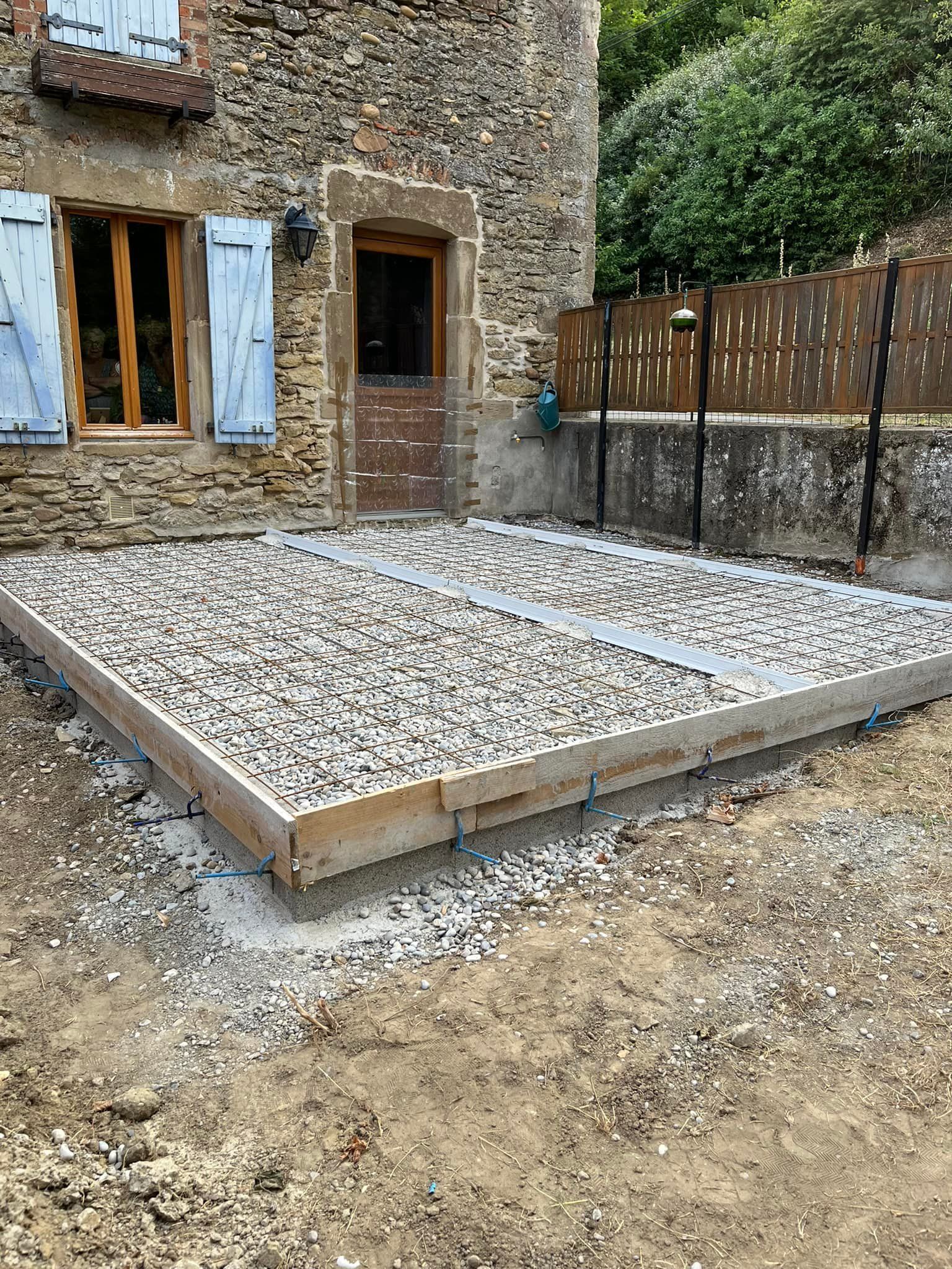 Une terrasse en gravier en construction jouxte un bâtiment en pierre avec des volets bleus et une porte en bois.