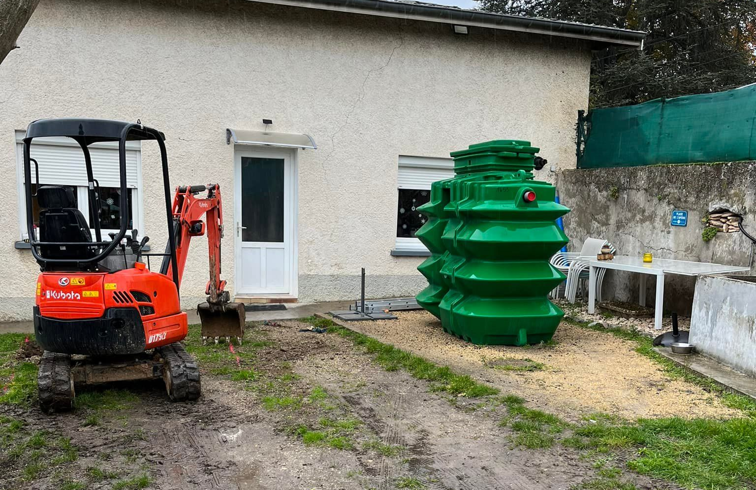 Mini-pelle orange et fosse septique verte près d'une maison. Chantier en cours.