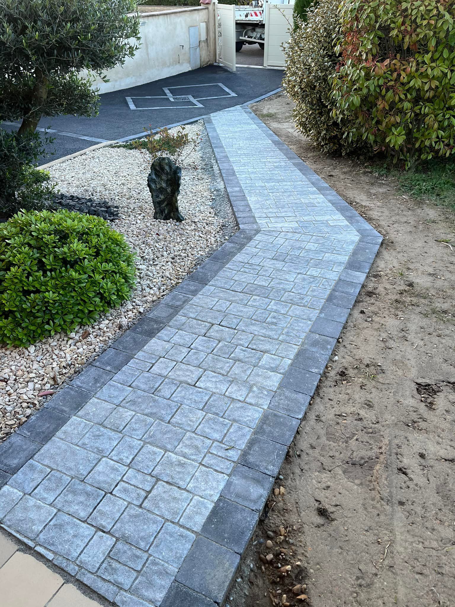 Allée en pierre bordée d'une frise, menant à une allée carrossable. Aménagement paysager : buissons, gravier et arbres.