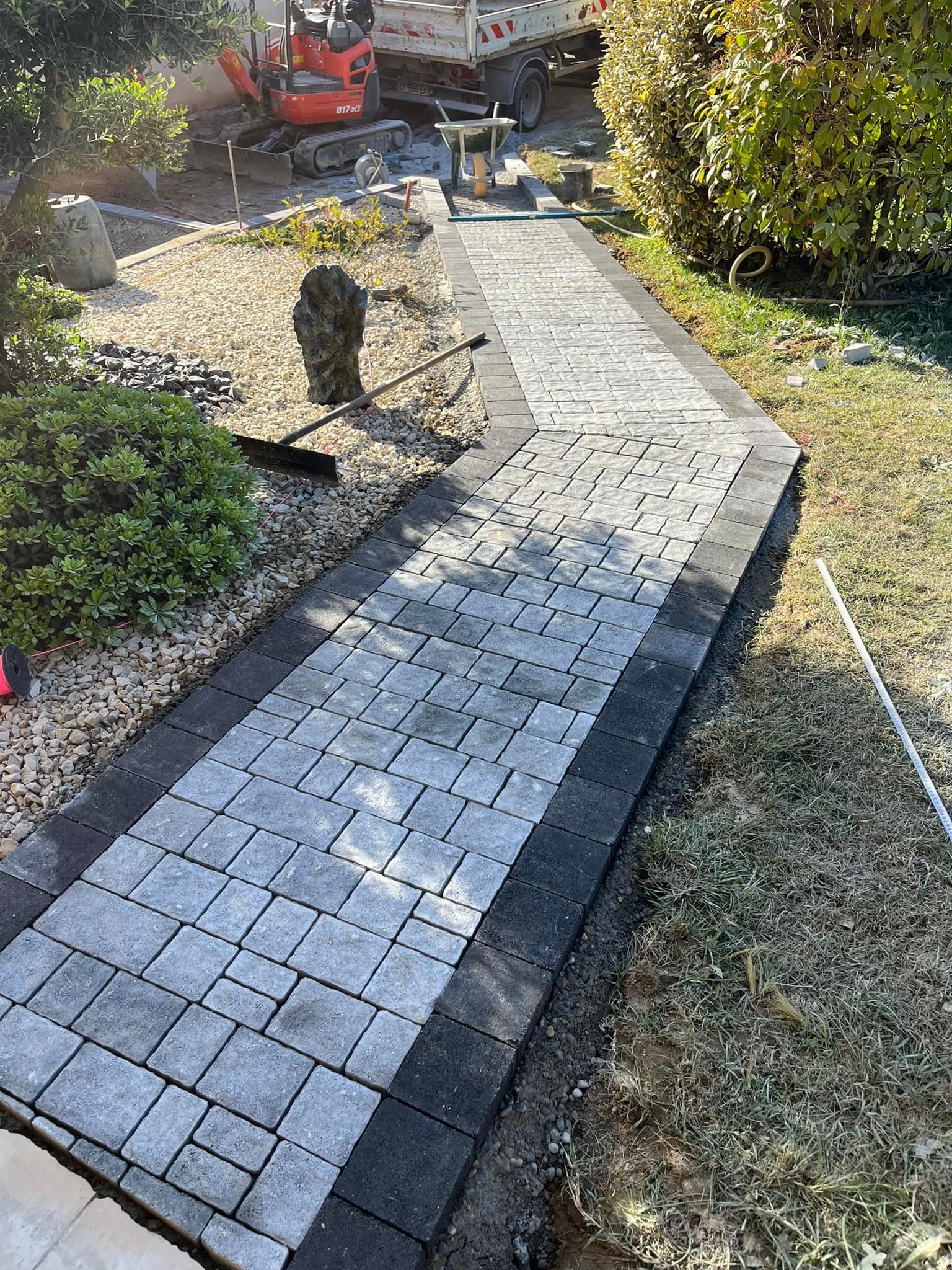 Allée en briques en construction, bordée de pavés foncés, de gravier et de gazon. Machines en arrière-plan.