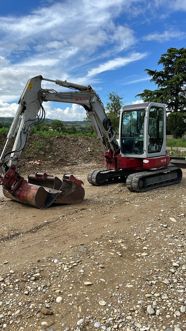 Une excavatrice rouge et blanche, godet déployé, sur une surface de gravier sous un ciel bleu.