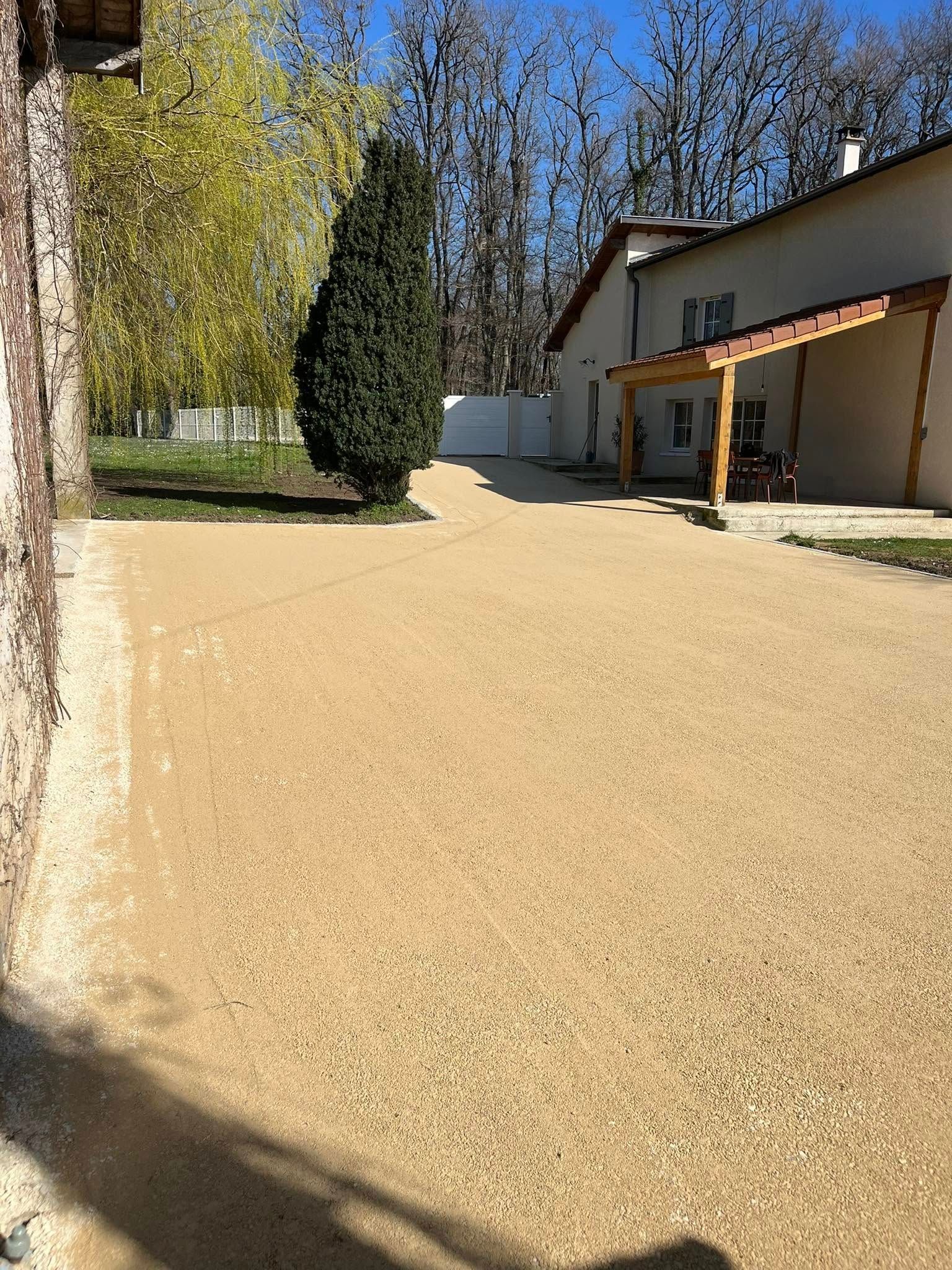 Une allée de gravier longe une maison avec un porche en bois et un grand conifère sous un ciel bleu.