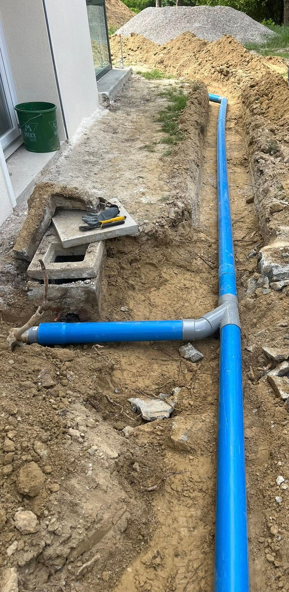 Des tuyaux bleus se trouvent dans une tranchée de chantier près d'un bâtiment. De la terre et des matériaux de construction entourent le tout.