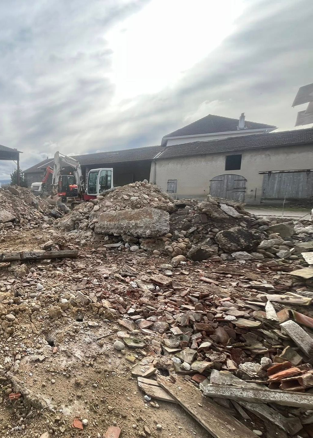 Chantier de démolition avec un tas de gravats au premier plan ; excavatrice et bâtiments visibles sous un ciel nuageux.