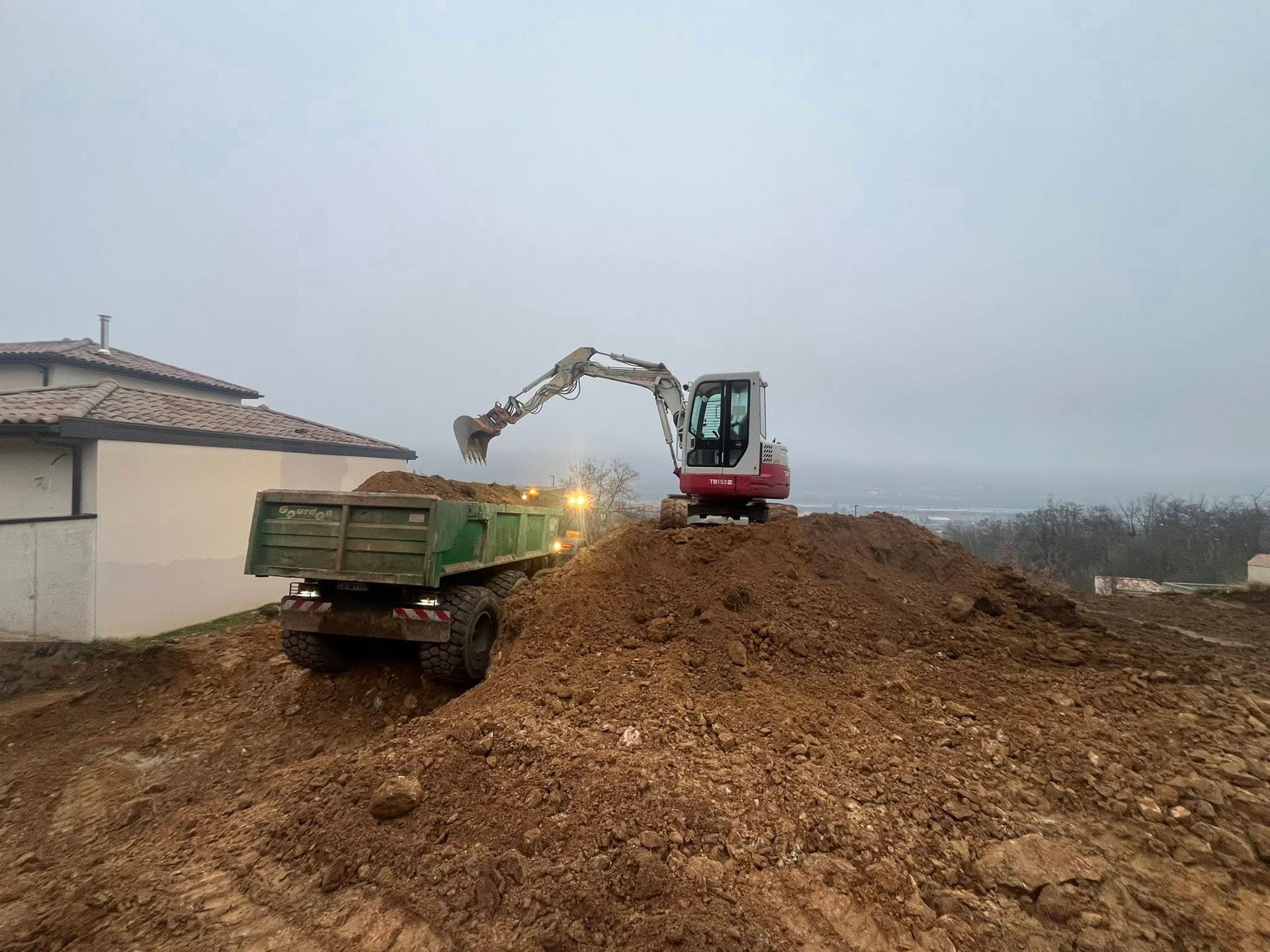 Une excavatrice charge de la terre dans un camion-benne, sur un chantier. Ciel couvert.
