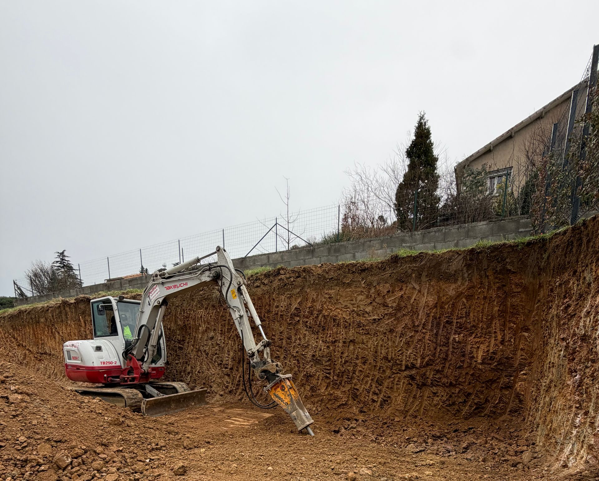 Une excavatrice creuse un mur de terre. En extérieur, sous un ciel nuageux, avec un bâtiment en arrière-plan.