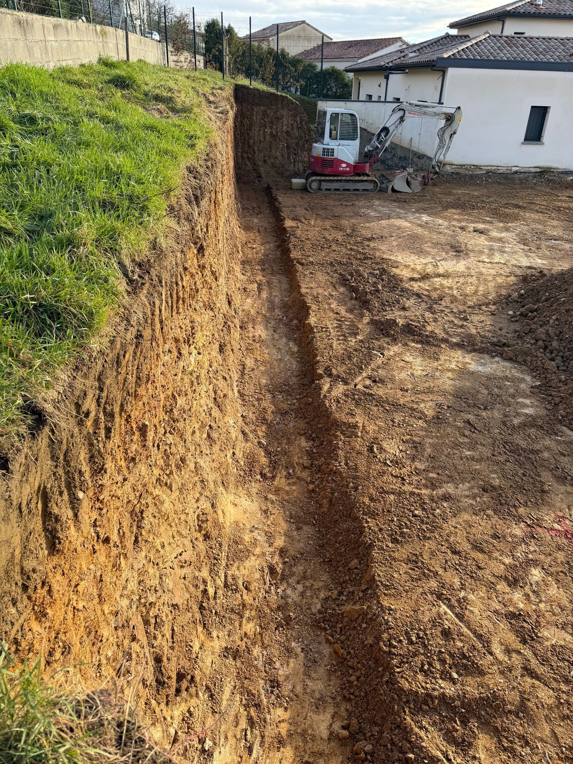 Tranchée d'excavation dans une cour avec une petite pelleteuse. Murs de terre, herbe verte en haut, maison blanche en arrière-plan.