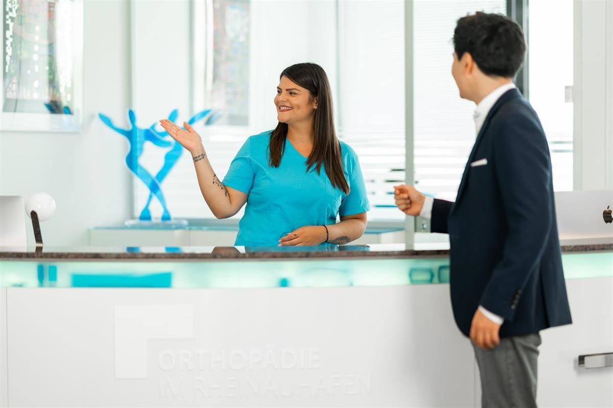 An einem Empfangstresen spricht eine Frau in einem blauen OP-Kittel mit einem Mann im Anzug.