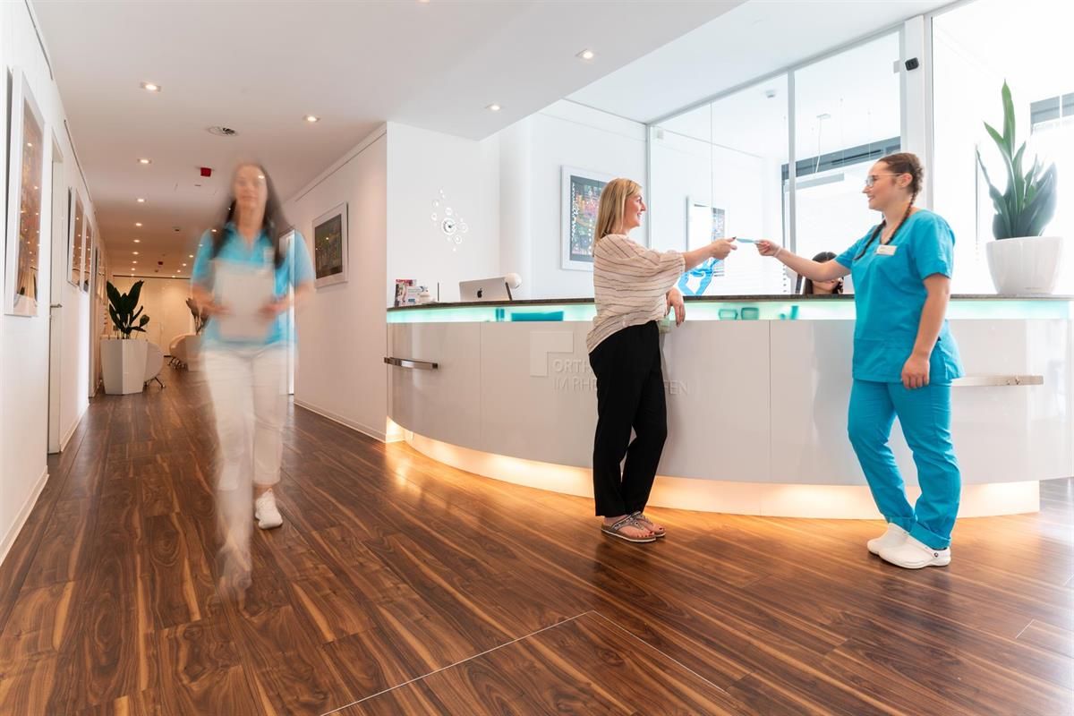 Eine Frau steht an einem Schalter in einem Krankenhaus und spricht mit einer Krankenschwester.