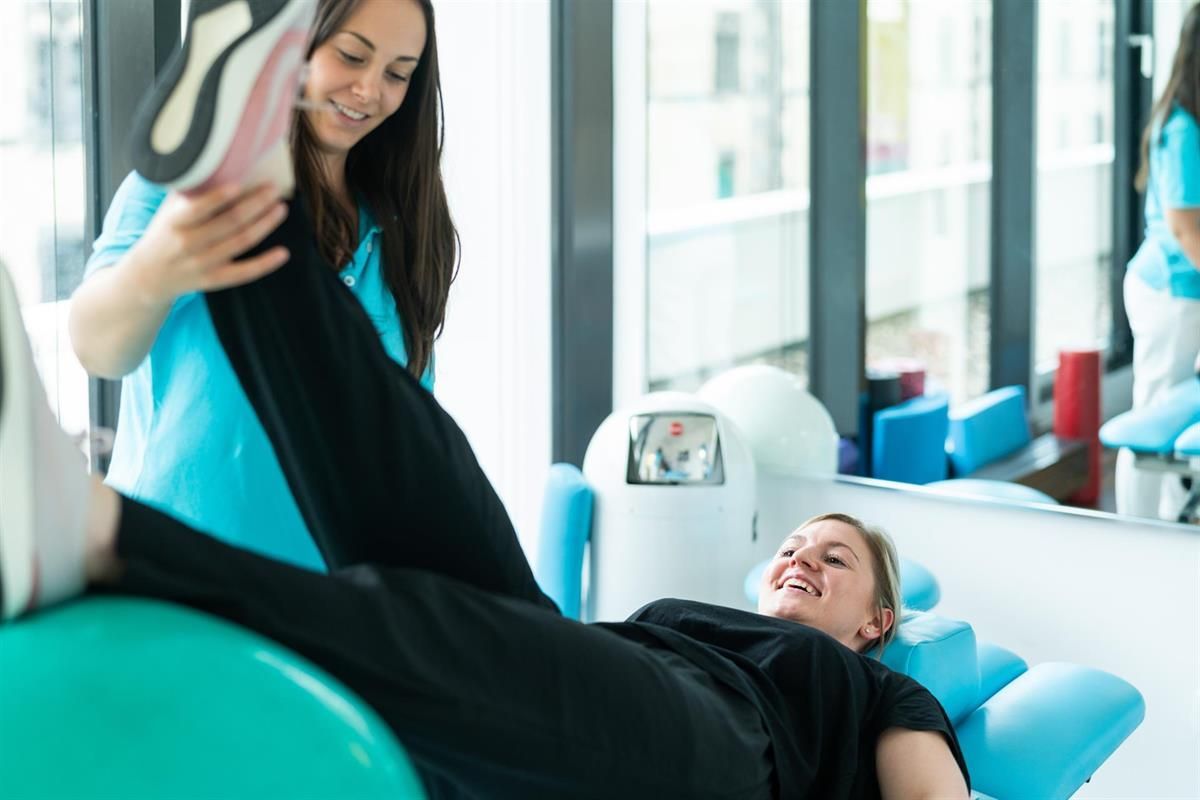 Eine Frau streckt ihre Beine auf einem Gymnastikball aus, während eine Krankenschwester zusieht.