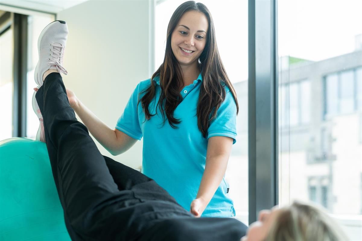 Eine Frau in einem blauen Hemd hilft einer anderen Frau , ihre Beine auf einem grünen Gymnastikball zu vertreten.