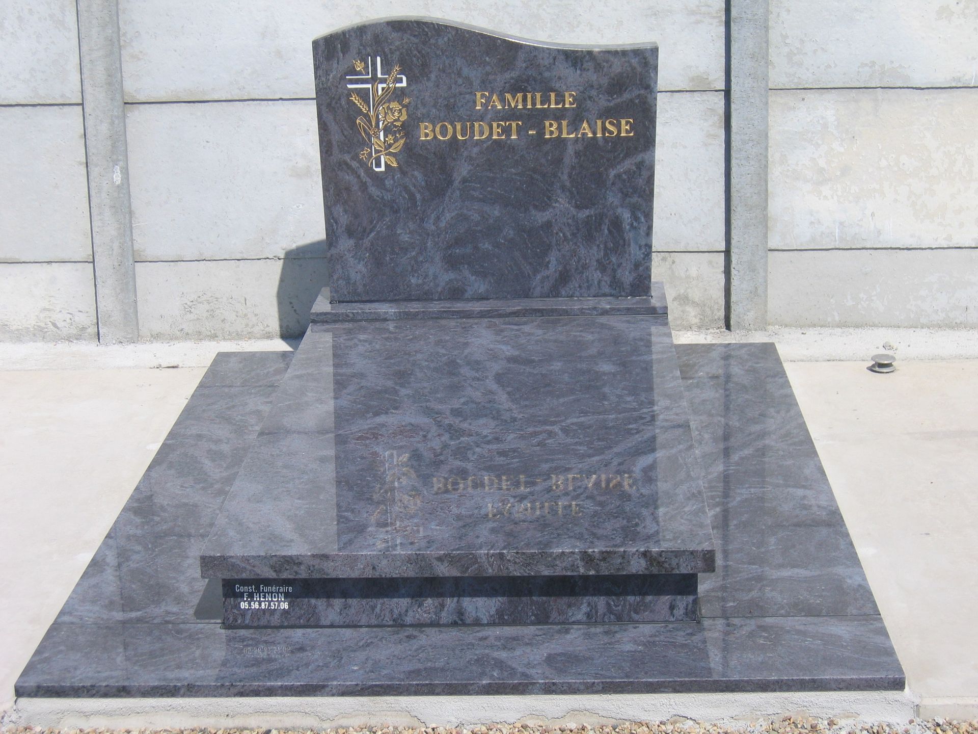 Pierre tombale en granit gris avec lettrage doré, croix et fleurs ; cadre extérieur.