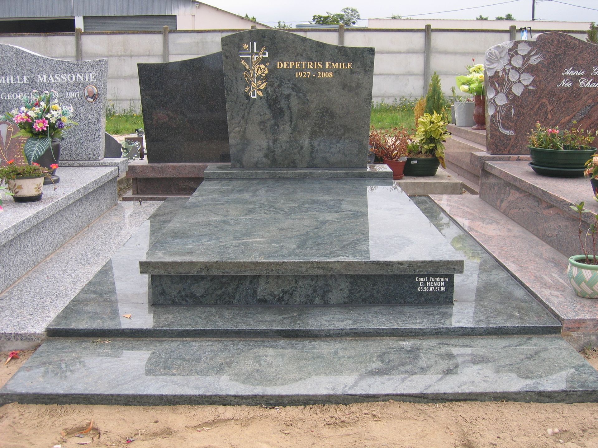 Une pierre tombale en granit avec des lettres dorées, une croix et des fleurs sur une tombe.