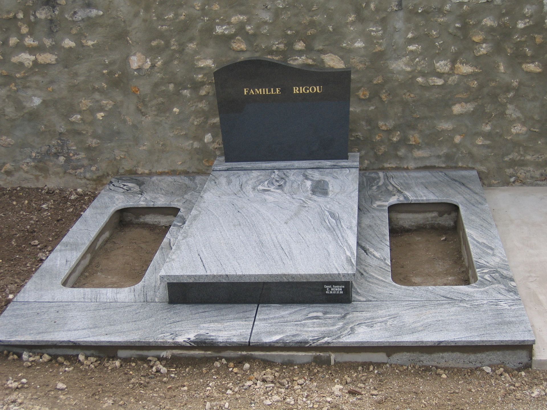 Tombe avec pierre tombale noire, socle en granit gris percé de deux ouvertures, contre un mur de pierre.