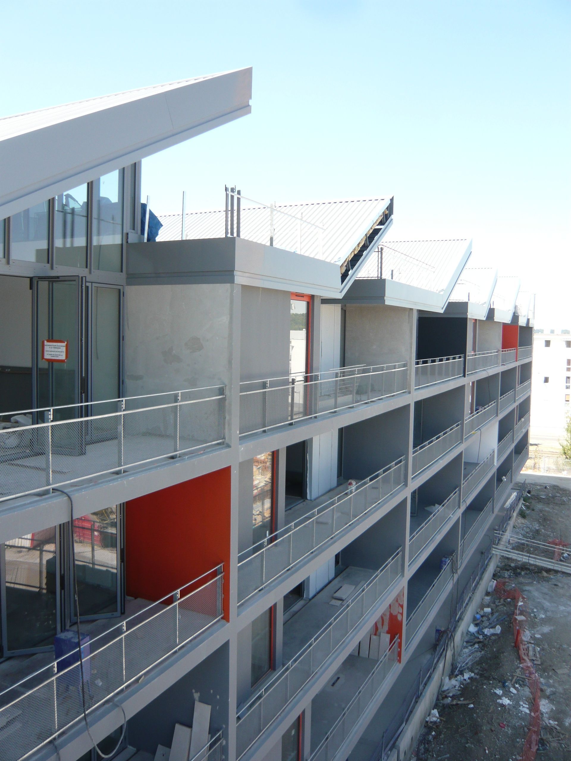 Bâtiment à plusieurs étages en construction avec béton gris, accents rouges et garde-corps en métal.