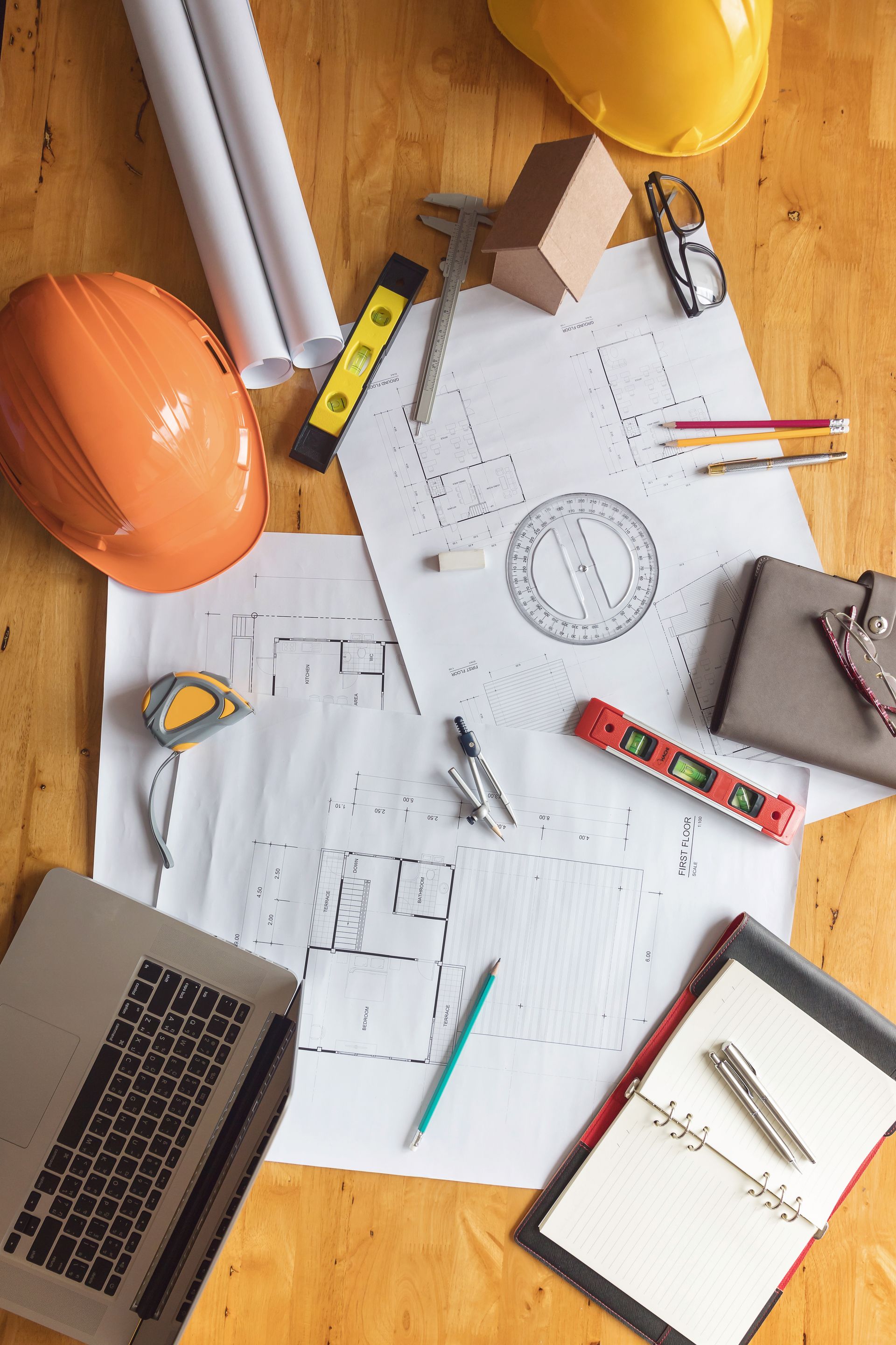 Espace de travail d'architecte avec plans, casques de sécurité, ordinateur portable et outils éparpillés sur une surface en bois.