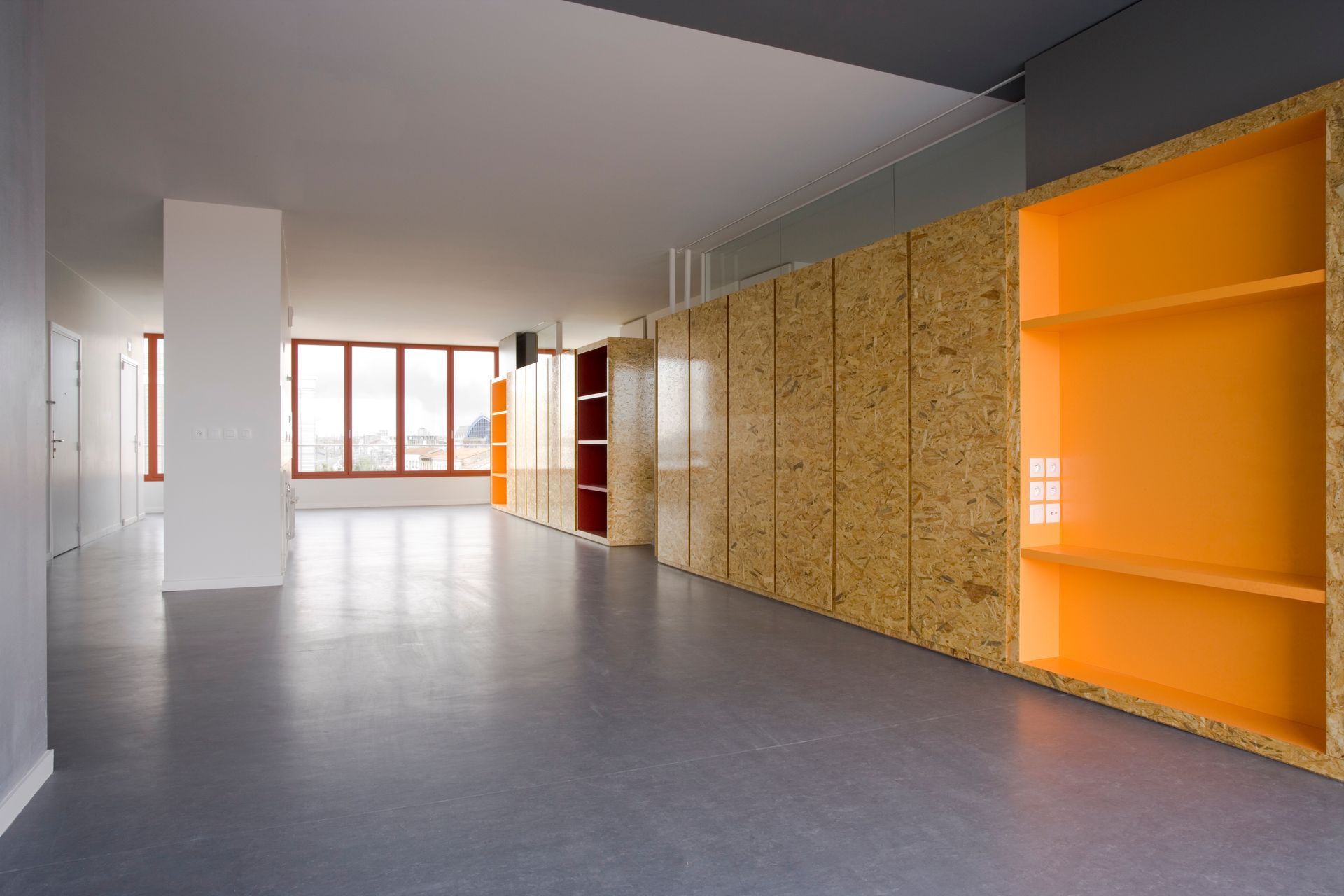 Pièce vide avec sol gris, lambris texturés et étagères orange. Fenêtres lumineuses au loin.