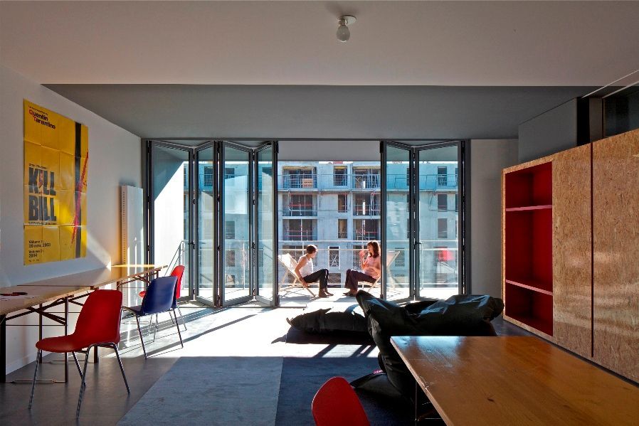 Intérieur avec portes vitrées ouvertes donnant sur un balcon où deux personnes sont assises. Chaises rouges et bleues, affiche jaune et mobilier en bois.