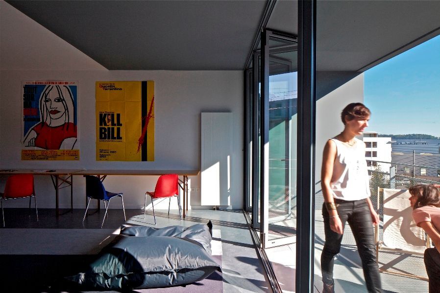 Pièce avec une paroi vitrée ouverte, une personne debout à côté. Affiches au mur, table avec chaises et poufs au sol.