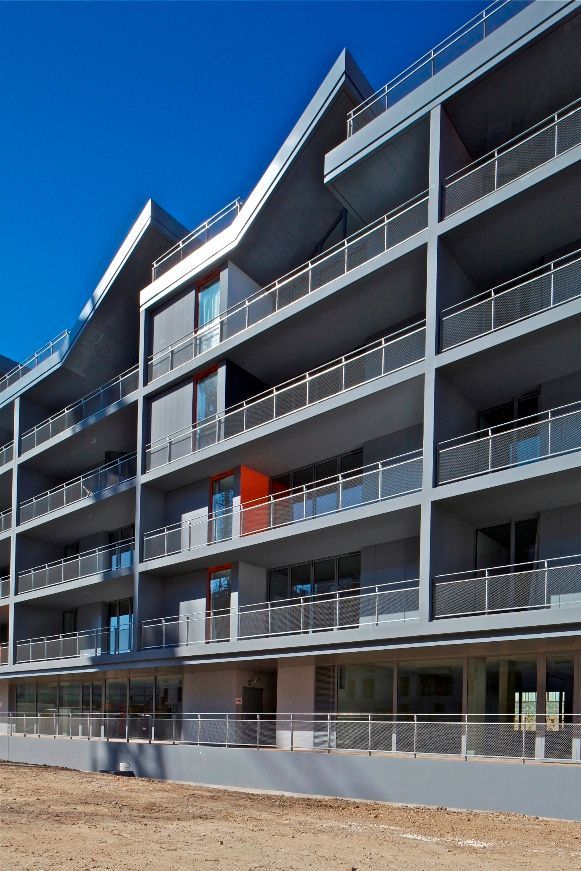 Bâtiment moderne gris avec balcons, balustrades argentées et accents rouges par une journée ensoleillée.
