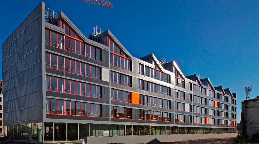 Bâtiment moderne gris et orange avec une ligne de toit en zigzag contre un ciel bleu clair.