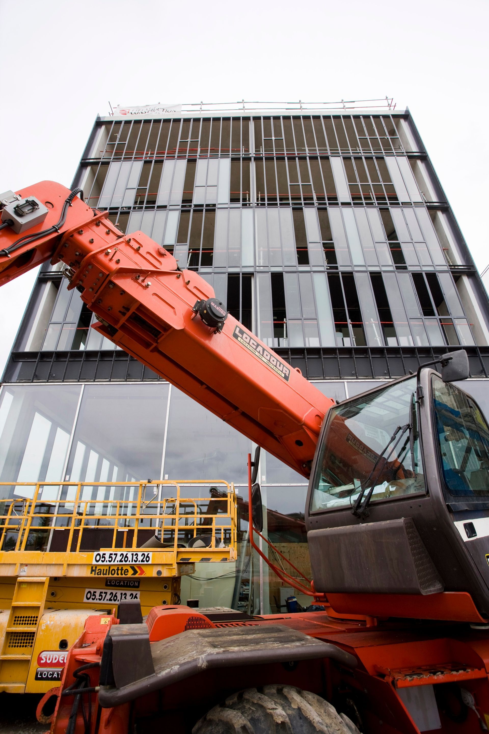 Chariot télescopique orange s'approchant d'un bâtiment moderne avec des panneaux de verre.