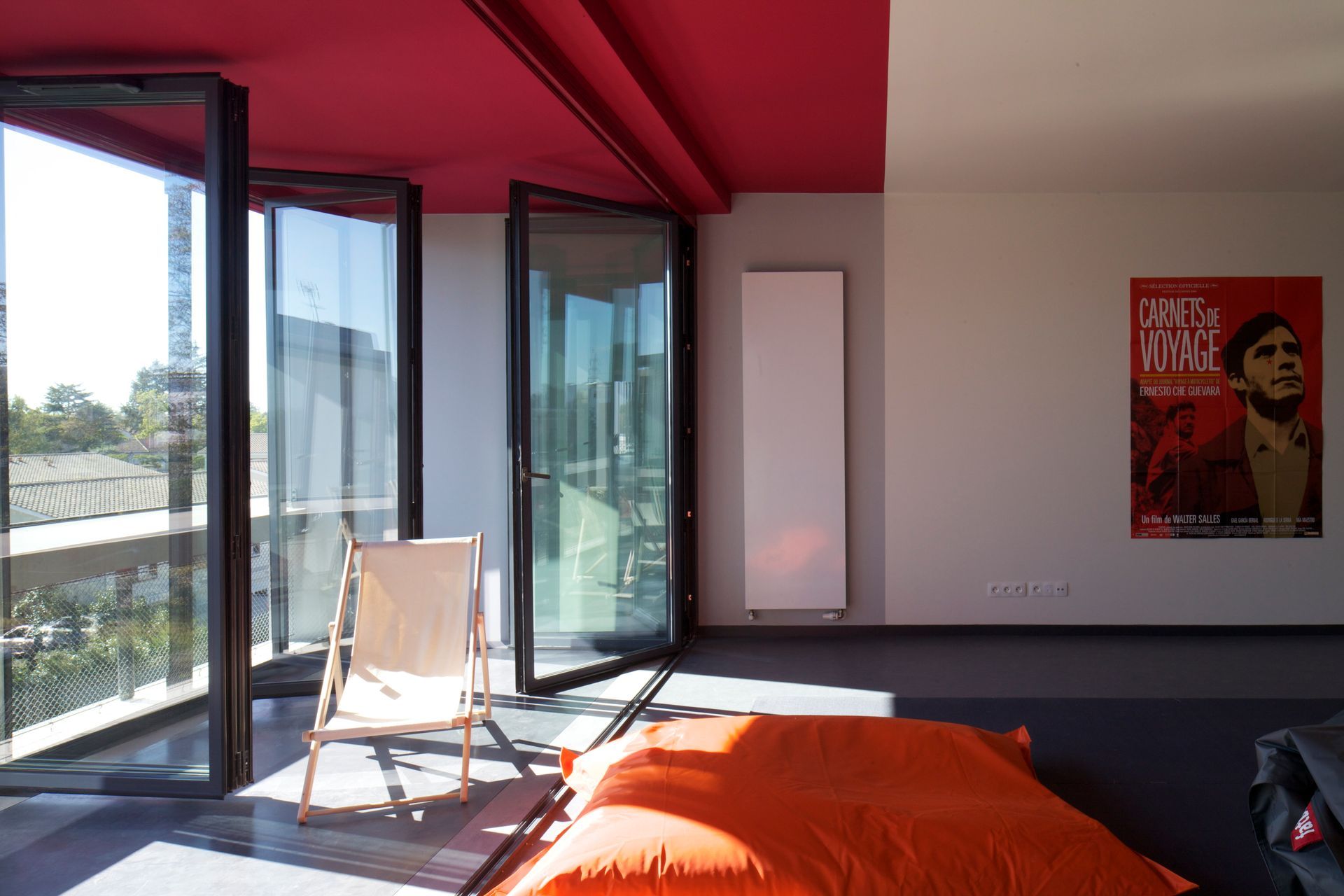 Chambre avec de grandes fenêtres, un plafond rouge, un pouf orange, une affiche et une chaise en bois.