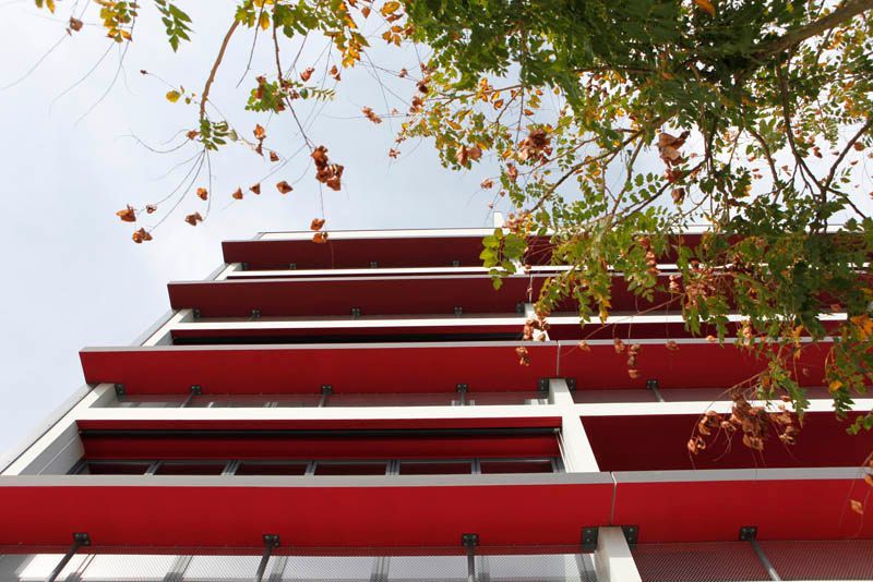 Extérieur d'un bâtiment moderne rouge et blanc avec des parasols rouges en surplomb, encadrés par des branches d'arbre.