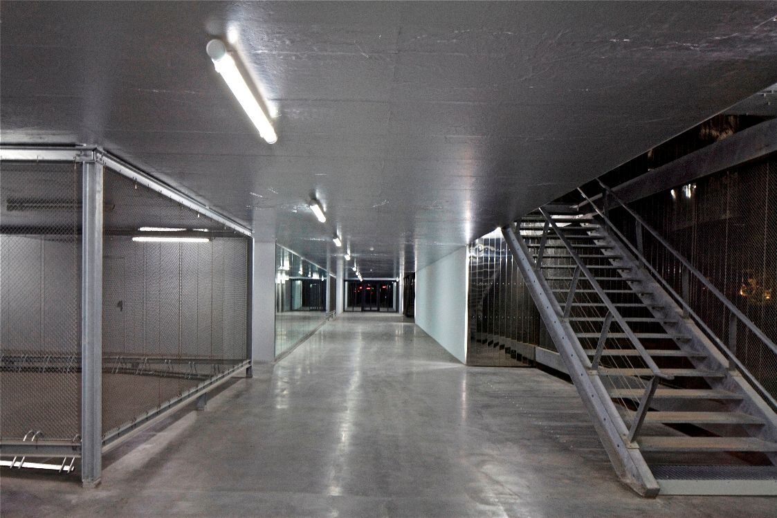 Un long couloir aux murs et plafonds métalliques, avec un escalier à droite. Des néons éclairent le sol en béton.