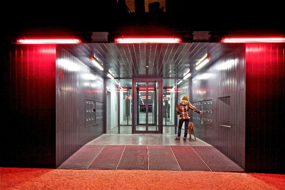 Entrée du couloir éclairée en rouge avec une personne et un chien. Une rampe métallique mène à un ascenseur vitré.