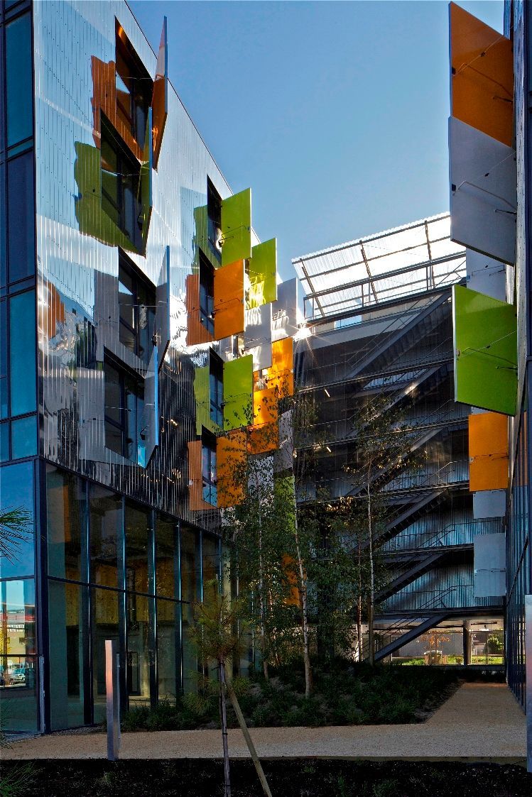 Bâtiment moderne avec panneaux colorés, façade en verre et cour arborée.